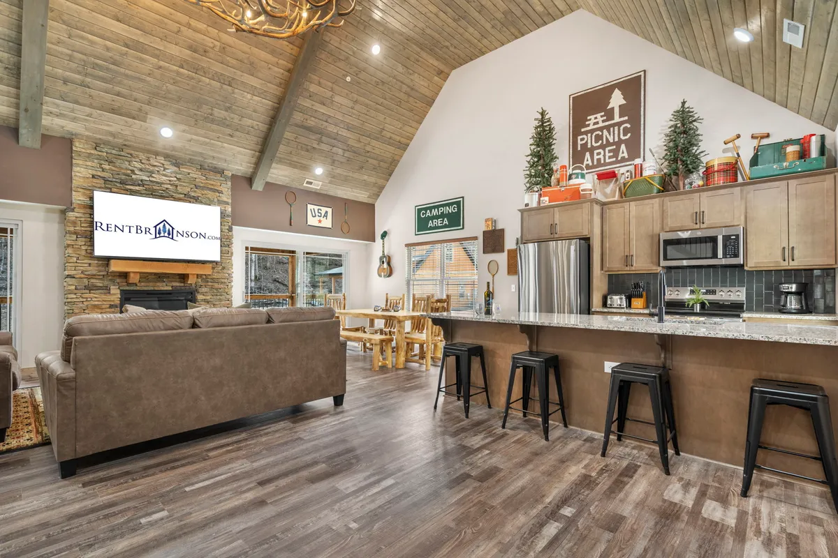 The image showcases a warm, open-concept living and dining area featuring a striking stone fireplace, expansive windows with natural light, a spacious wooden dining table, a gleaming granite countertop in the kitchen, and elegant chandeliers. The space exudes a cozy yet upscale cabin feel, perfect for family gatherings, game nights, and relaxing by the fire.