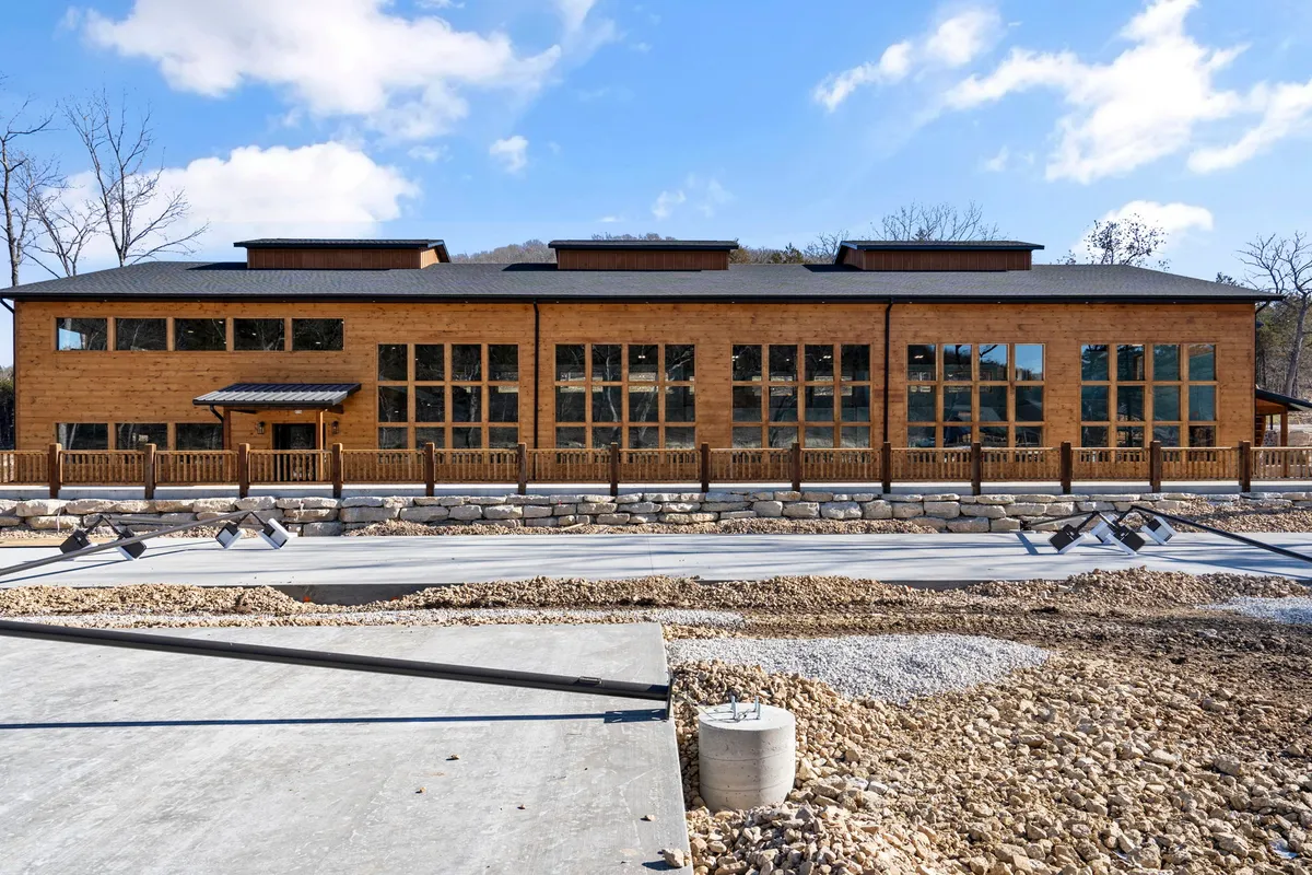 This exterior image showcases the inviting wooden cabin with large windows, a stone base, and landscaped surroundings. The scene hints at communal gatherings, outdoor activities, and the warmth of shared experiences—echoed by the sunset glow and the patio area ready for shared meals or evening bonfires.