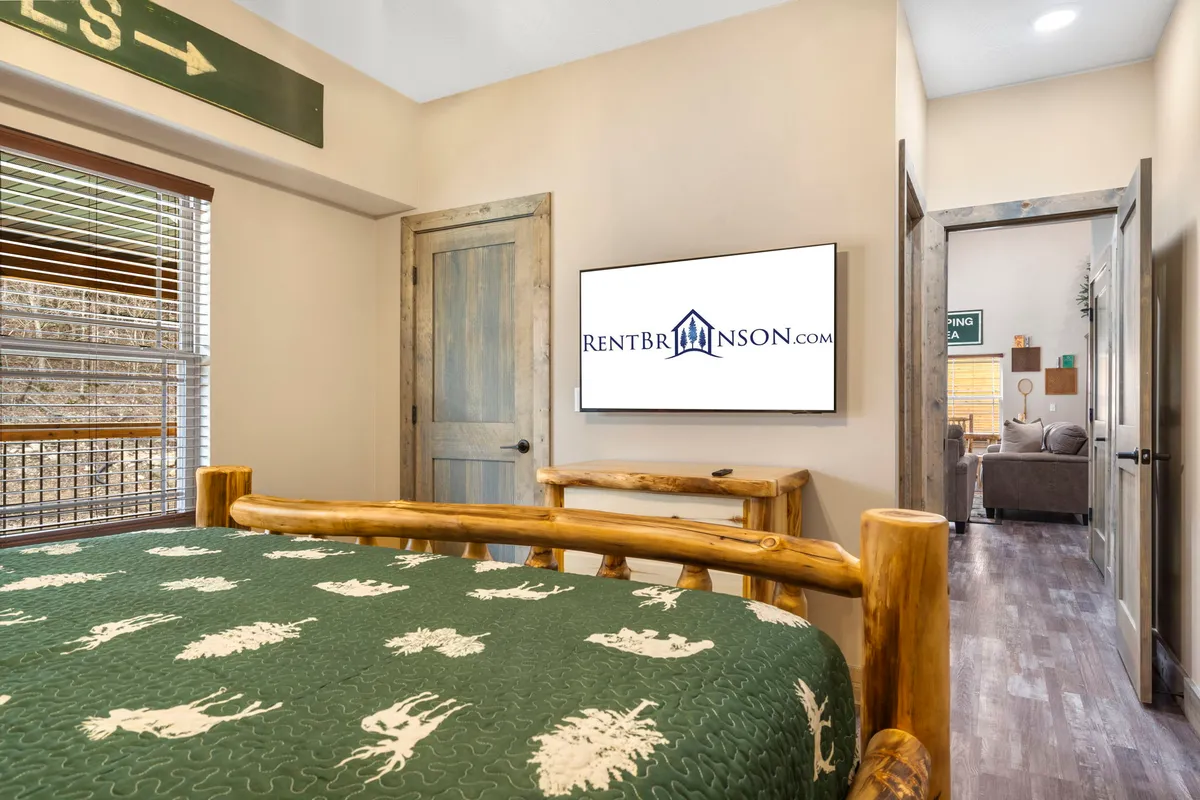 A welcoming bedroom with a sturdy log bed frame, a television for entertainment, a wooden door, and a window with blinds allowing glimpses of the outdoors. The setting is restful and inviting, ideal for curling up at the end of the day.