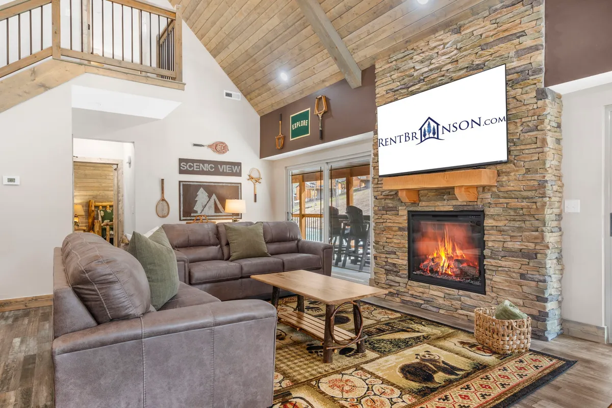 A welcoming living room featuring a striking stone fireplace as the focal point, surrounded by a comfortable sofa, a large television, and a wooden coffee table. The furniture is arranged to promote conversation and relaxation, perfect for gatherings. Warm tones and natural materials create an inviting, homey atmosphere ideal for family game nights or cozy evenings by the fire.