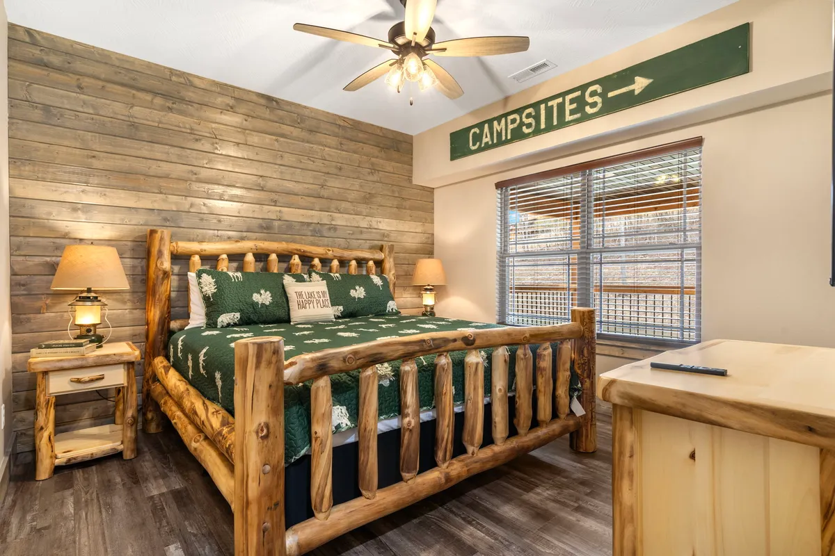 A cozy bedroom featuring a rustic log bed with a green blanket, warm wooden walls, a 'campsites' sign, and soft table lamps that create a tranquil, inviting atmosphere. The space feels serene and perfect for unwinding, with natural light filtering in.