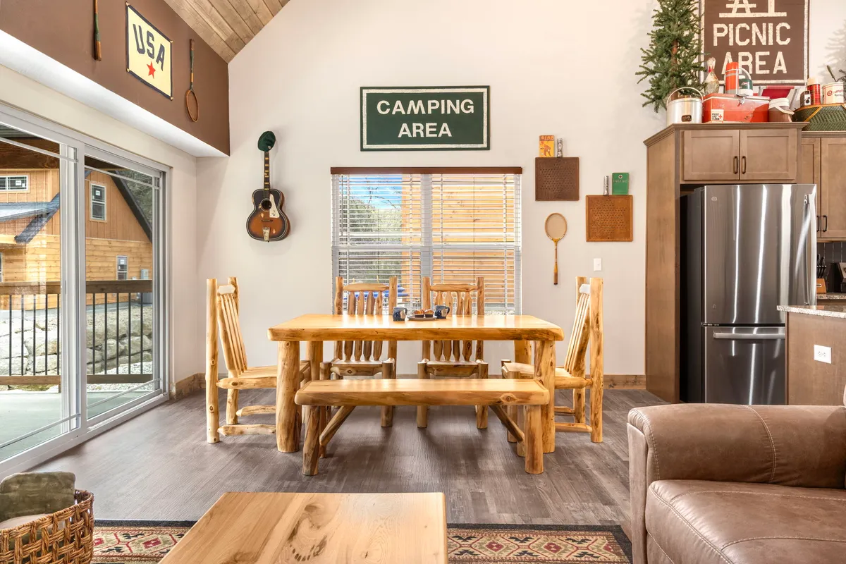 A spacious dining area that features a large wooden dining table, wall-mounted guitar, and camping-themed signage, evoking a relaxed, communal feel. The table is large enough to accommodate groups, and the decor emphasizes sharing meals, music, and stories in a casual setting. The open design hints at easy movement between spaces.