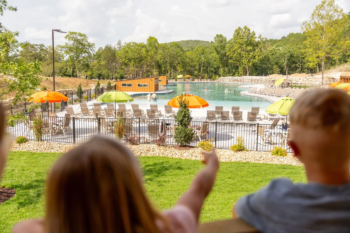 An exterior view of the outdoor swimming pool area, featuring colorful umbrellas, lounge chairs, and children joyfully splashing in the water. The setting is surrounded by trees, creating a cheerful and active atmosphere perfect for summer fun.