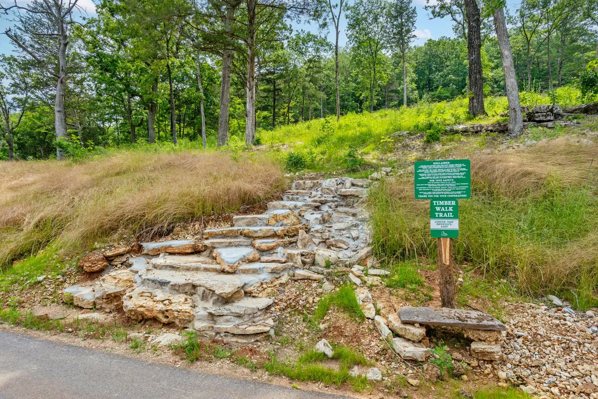 A rustic trail sign marks the beginning of a stone path surrounded by dense greenery, inviting visitors to embark on a hike or nature walk through the Ozark landscape. The untouched vegetation and gentle sunlight evoke a sense of peaceful exploration.