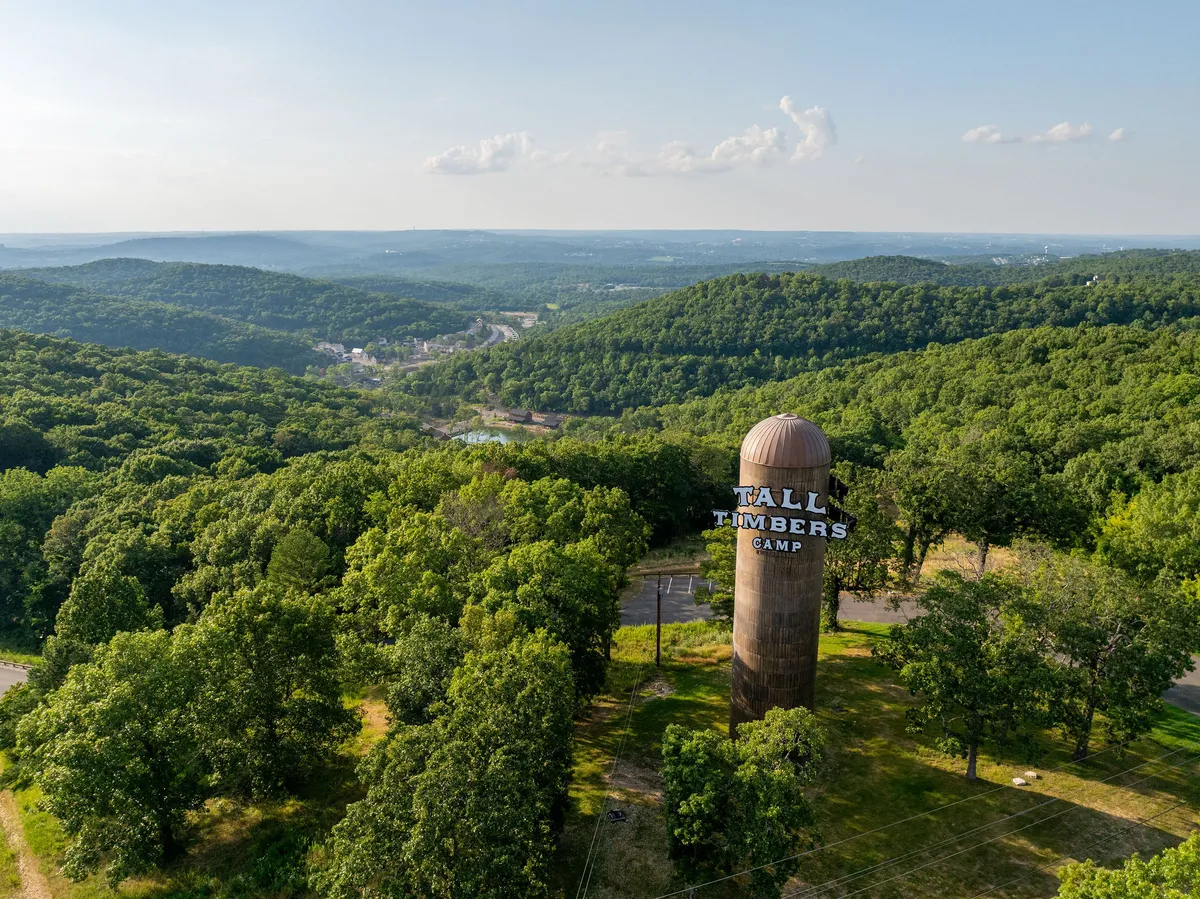 A striking water tower emblazoned with 'Tall Timbers Camp' rises above tree-lined rolling hills, immediately conveying the sense of a newly established, adventurous resort community. The setting sun casts a warm glow over the expansive landscape, hinting at both ongoing development and the promise of memorable outdoor experiences.