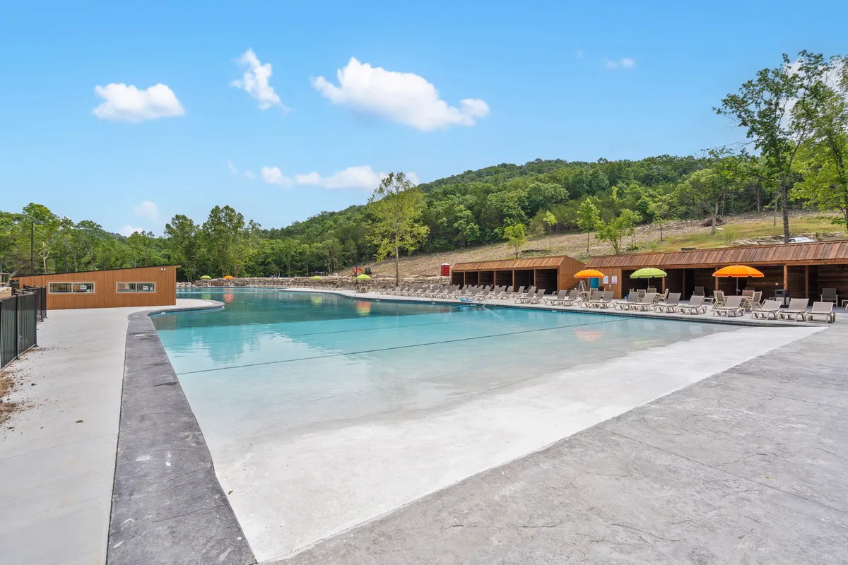 A sparkling outdoor swimming pool bordered by lounge chairs and vibrant umbrellas, nestled among tall trees. Children laugh and play in the water, while families enjoy poolside barbecues and relaxed sunbathing, all set against a backdrop of ongoing freshness and potential development.