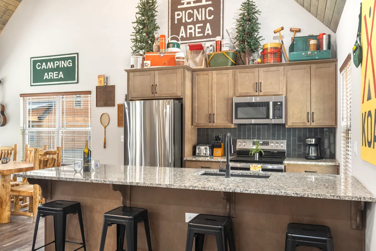 A cozy, rustic kitchen scene with a granite countertop, stainless steel appliances, and a wooden dining table. Decorative picnic and camping area signs evoke an inviting, playful atmosphere, while the kitchen's openness suggests a space for cooking together and hosting group meals. The design balances functionality and comfort.