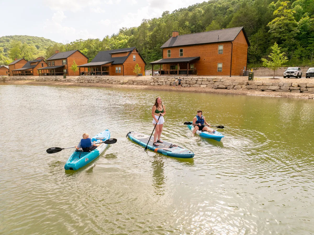 Three kayakers glide across a calm water body, with charming wooden cabins nestled along the shoreline and lush green hills rising in the background. The scene is alive with activity and natural beauty, hinting at the variety of outdoor adventures available.